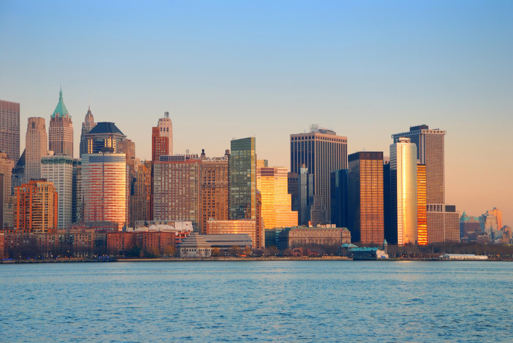 A photograph capturing the warm glow of sunset reflecting off the high-rise buildings of Lower Manhattan's skyline, as viewed from across the water, from the Staten Island Ferry.